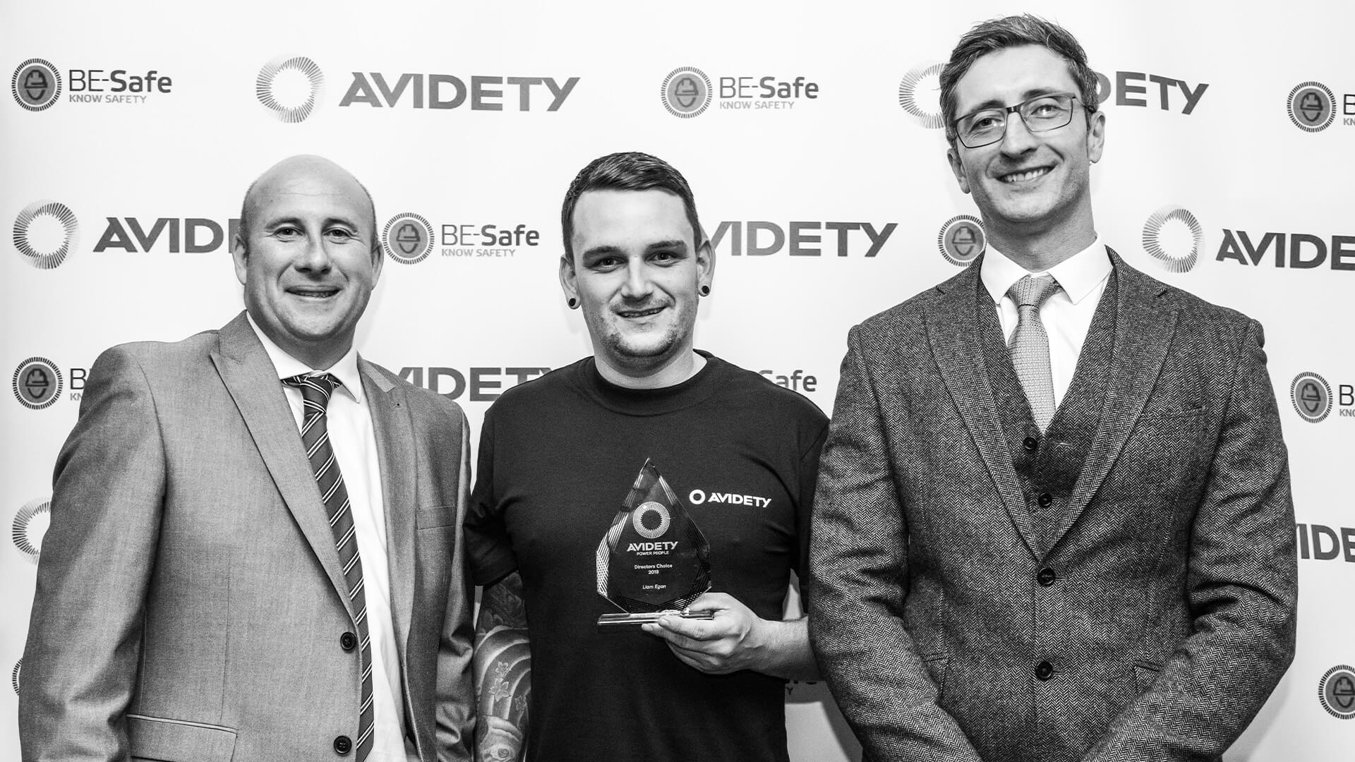 3 Men Standing in front of Avidety Logo, Middle man Holding Trophy