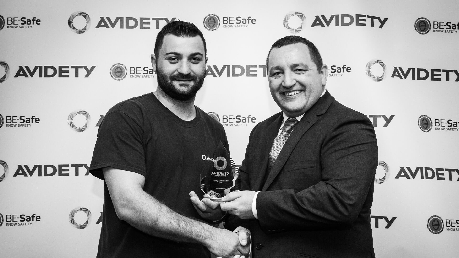 2 Men Standing in Front of Avidety Logo Shaking Hands and Holding a Trophy, the Photo is in Black and White